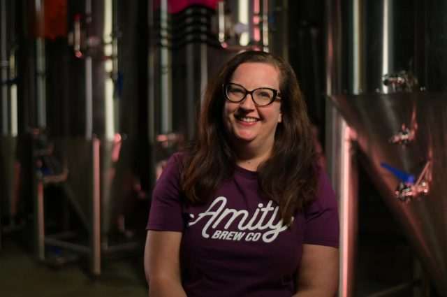 A photo of the Amity Brew Co communications manager in a branded T shirt sitting in front of their brewing machinery