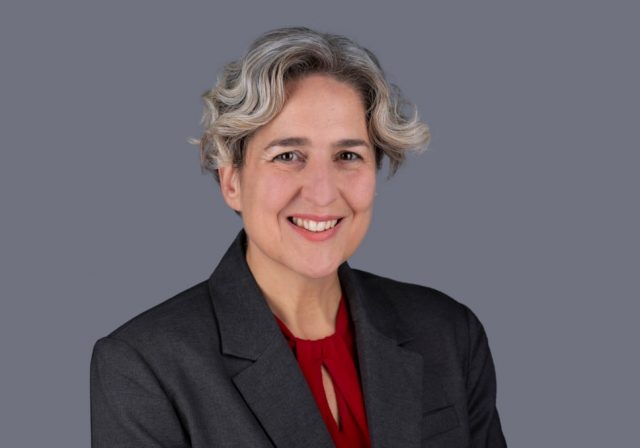 A headshot photo of the Mayor of Hackney, Caroline Woodley. She is a middle aged woman with wavy grey hair, wearing a grey suit jacket over a red blouse.