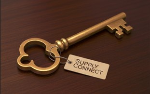 A thick brass key is lying on a polished dark wooden surface. The key is slightly ornate, implying it opens the grand doors. A tag is tied to it which says SUPPLY CONNECT.