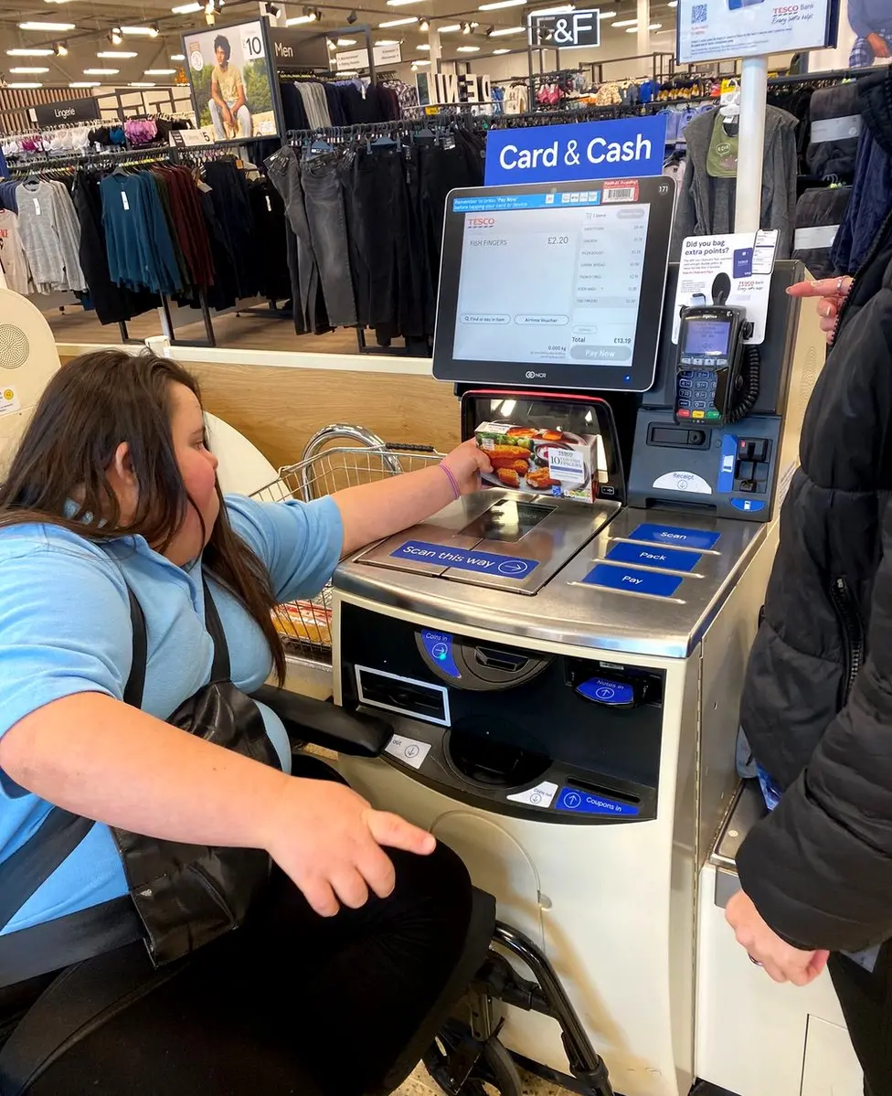 A shopper in a wheelchair tries to hold her bag on her lap as she reaches to scan a ready meal at a Tescos self-service till.